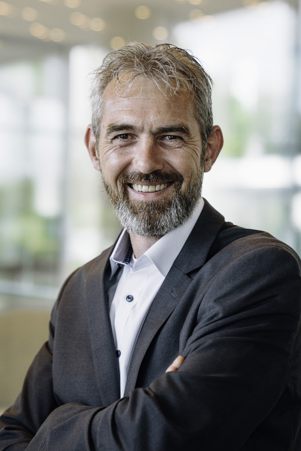 Das Bild zeigt Marco Brentführer, Ihren Ansprechpartner bei Solarlux. Er ist ein freundlicher, selbstbewusst lächelnder Mann mit grauem Bart und kurzen Haaren. Er trägt einen dunklen Anzug mit einem weißen Hemd. Der Hintergrund ist modern und lichtdurchflutet. Sein professionelles Auftreten vermittelt Kompetenz und Offenheit.